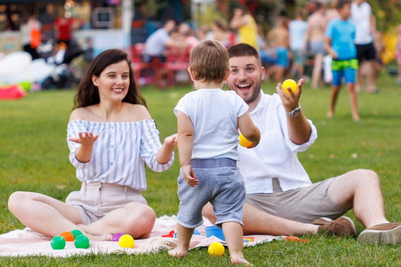 Playful cute little boy running towards his parents while having picnic in the park; mother and father sitting on picnic blanket having fun playing with their son