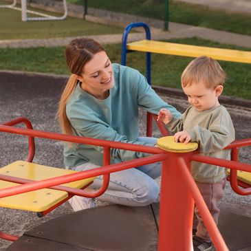 A woman and toddler playing on a playground merry-go-round.