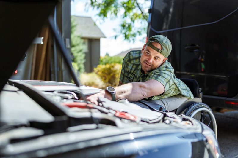 An adult Caucasian man who has a physical disability and uses a wheelchair works on his car at home in the garage on a beautiful Autumn day.