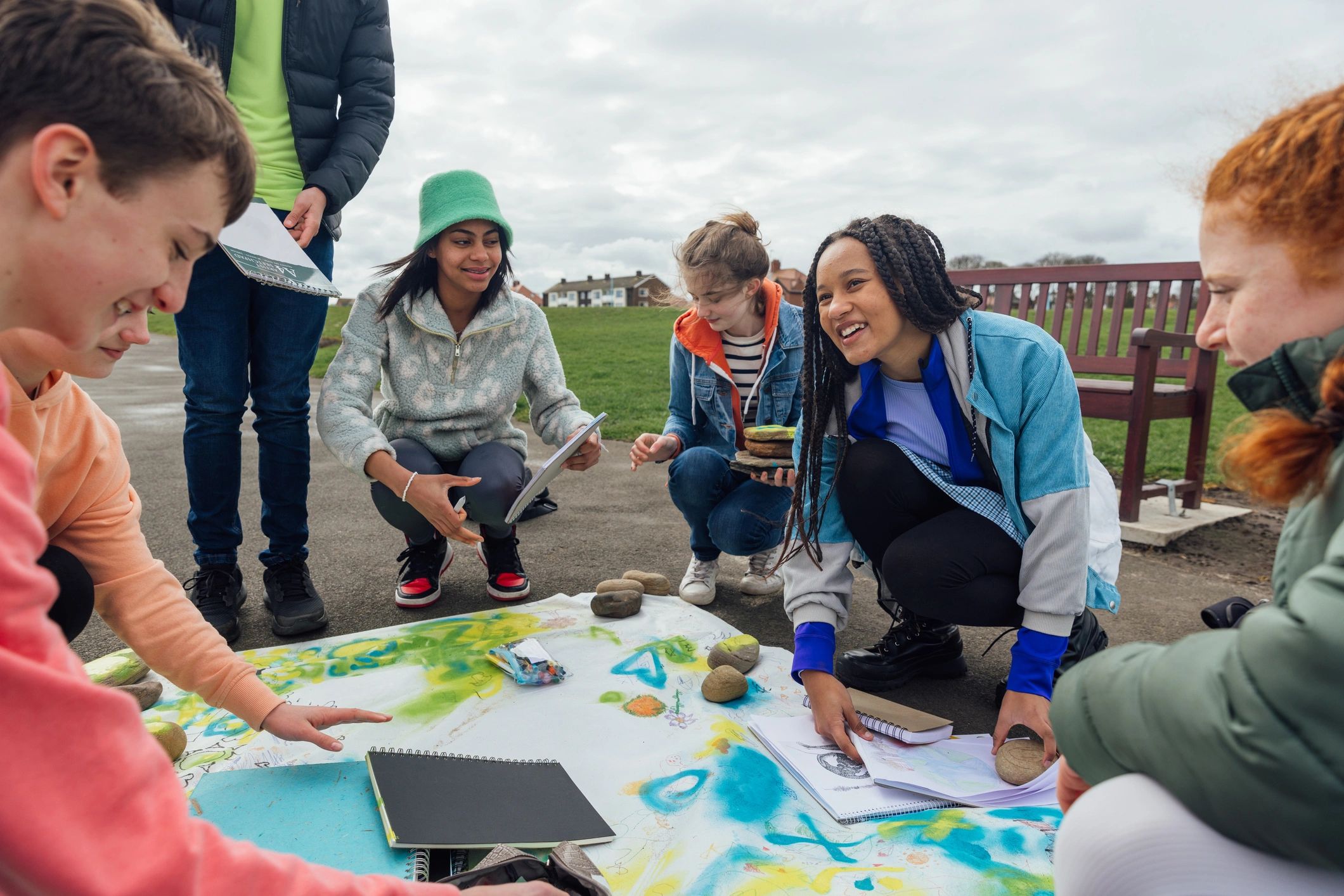 Group of smiling teens painting on a large sheet outdoors.