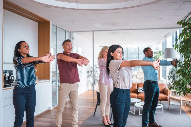 A group of cheerful office employees, diverse in ethnicity with males and females, doing stretching exercises together in a well-lit corporate office setting, promoting workplace wellness and teamwork.