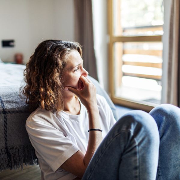A woman in casual clothes sits on the floor, looking thoughtfully out a window.