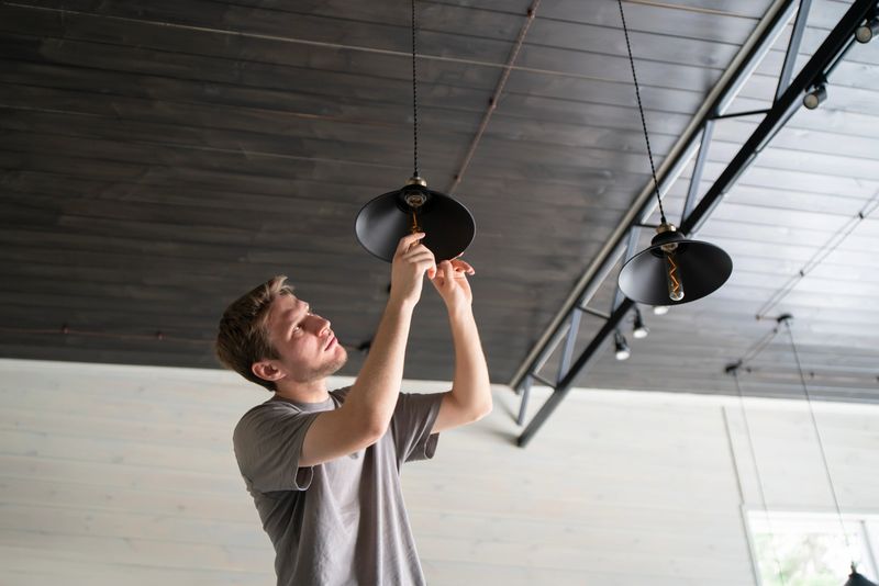 A man is captured in the act of changing a light bulb in a hanging ceiling light fixture, with natural daylight illuminating the scene.