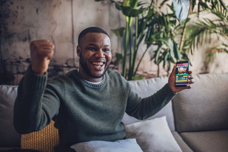 Jubilant man with phone in hand celebrates winning a sports bet, the betting app's screen reflecting his success.