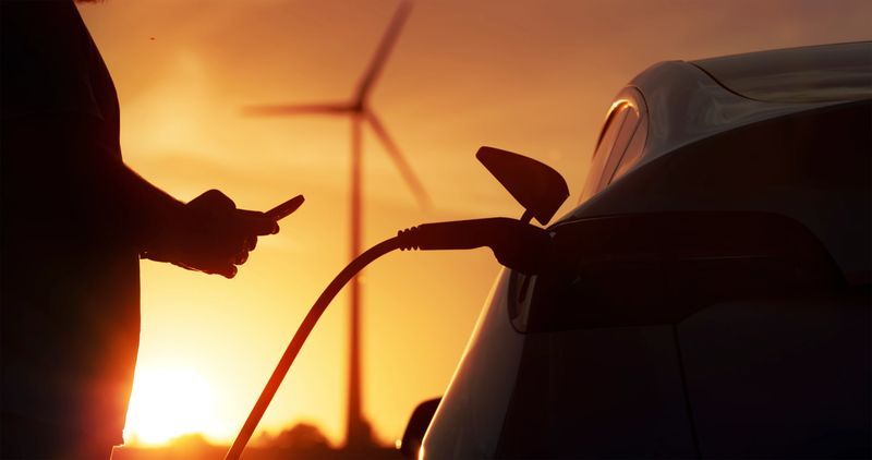 Electric car being charged with plug-in power cord at charging station during sunset.