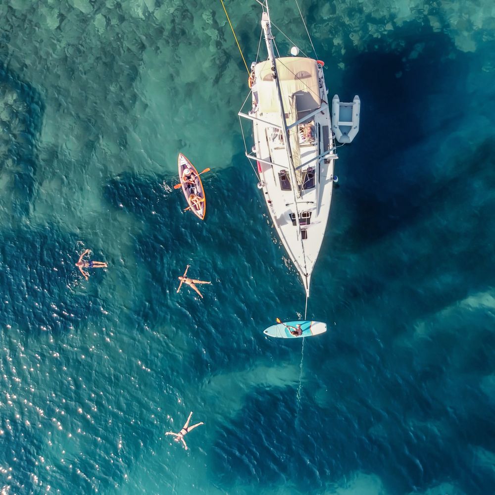 Aerial view of people enjoying water activities around a anchored sailboat in clear blue sea.
