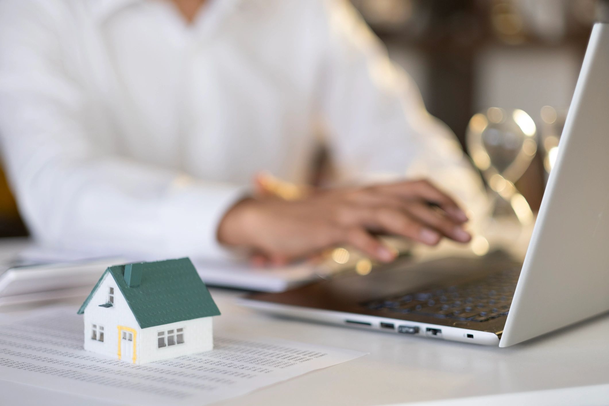 Small house model on papers with person working on laptop in background.