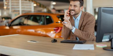 Car salesman talking on phone at dealership desk with orange car in background.