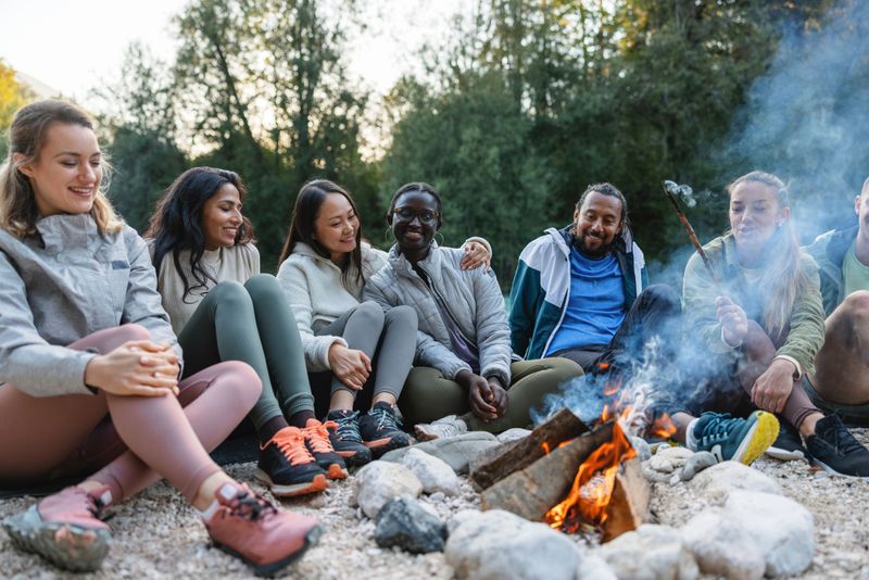 A mixed ethnic group of friends laughing and relaxing around a campfire in a natural outdoor setting.