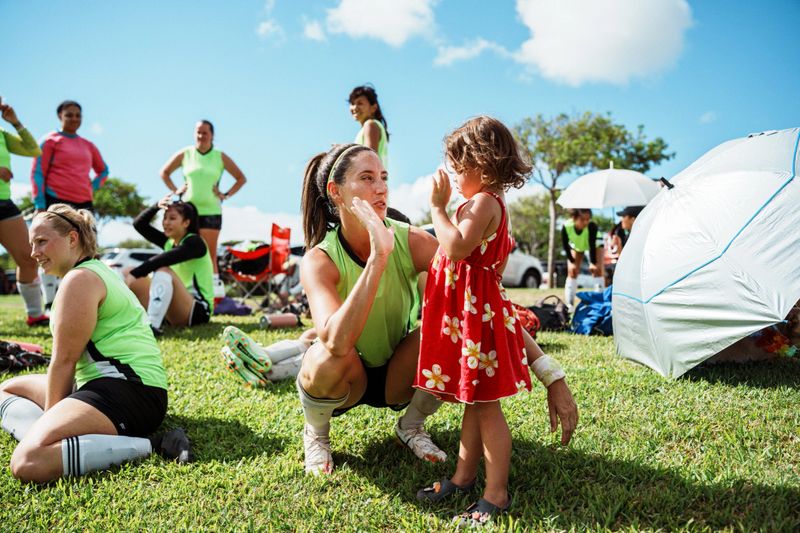 A beautiful and fit female athlete playing in a club soccer tournament in Hawaii gives her three year old daughter a high-five on the sidelines of the field after the game.