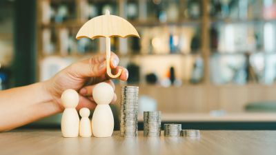 Hand holding umbrella over wooden figures and stacked coins.