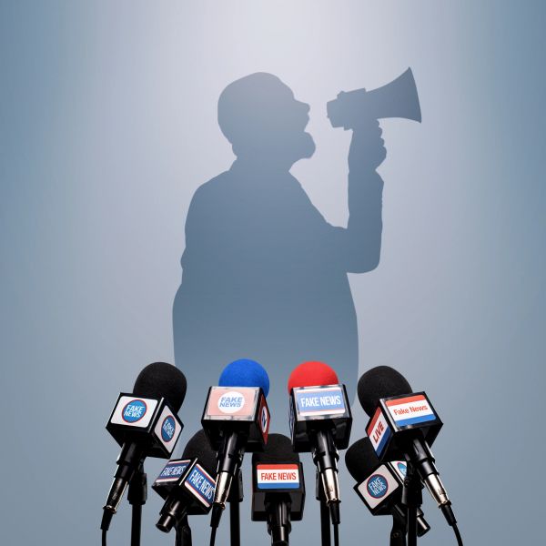 Shadowed figure shouting into a megaphone behind microphones labeled 'Fake News'.