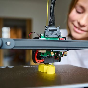 A girl watching a 3D printer creating a yellow object.