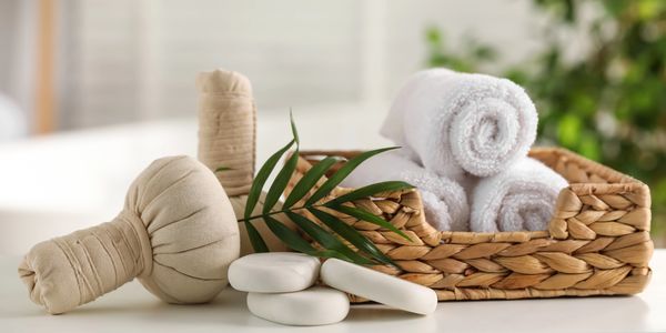 Rolled white towels and spa essentials arranged in a woven basket with green leaves.