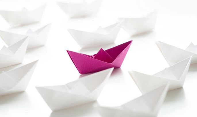 A single purple origami boat stands out among many white boats.