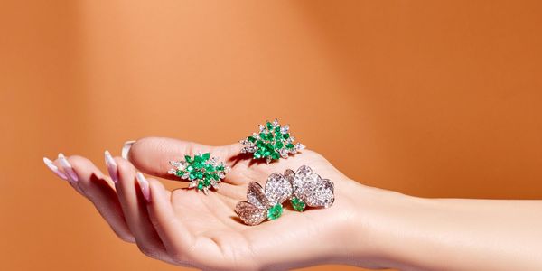 Hand holding sparkling green and clear gemstone earrings against a warm brown background.