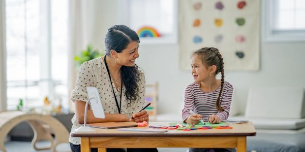 Teacher and young girl smiling while doing crafts at a table in a classroom.