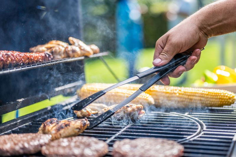 Chicken wings and corn being roasted on grill and flipped with tongs close up