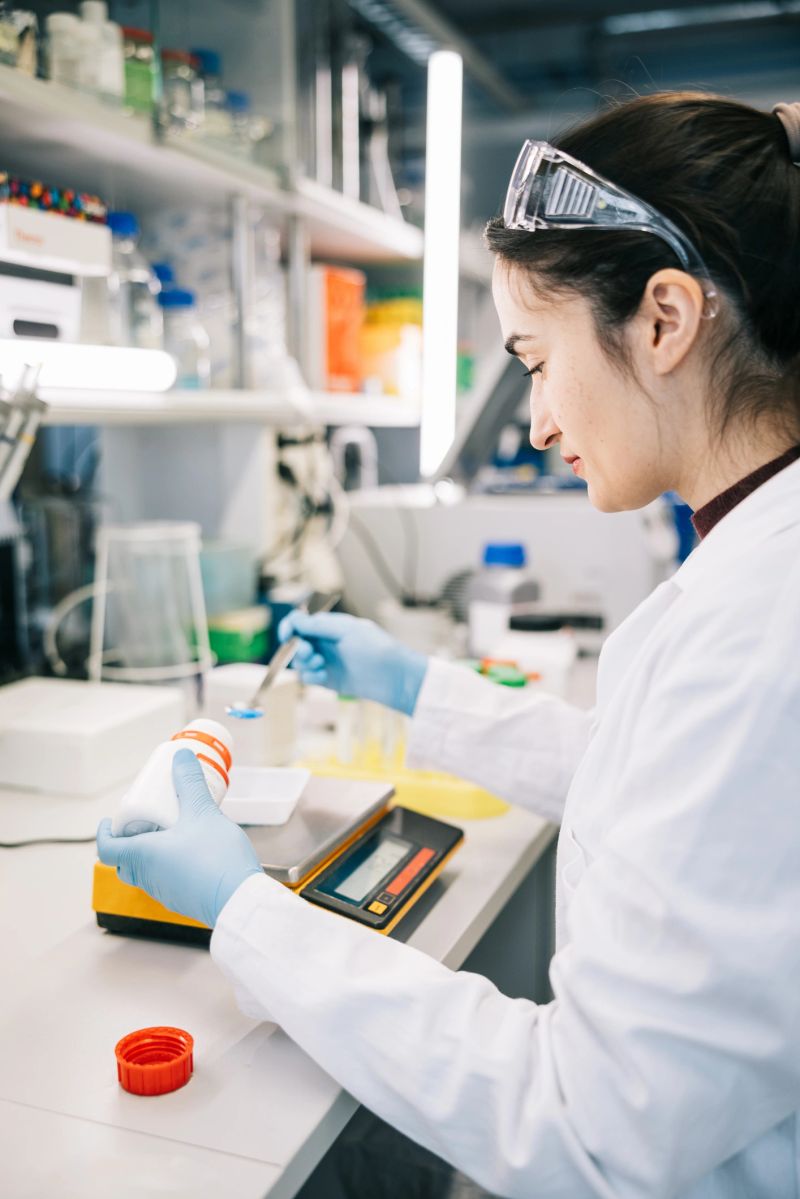 A dedicated female scientist, in a white lab coat, focused on weighing chemicals with precision on a digital scale in a well-equipped laboratory