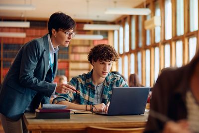 Teacher helping student with laptop in library.