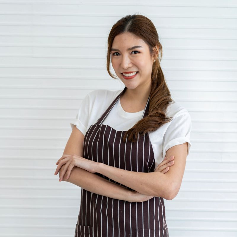 Portrait of cheerful young Asian woman working as barista, smiling with her arms crossed while wearing white t-shirt and apron. She stands against a pure white background wall inside the cafe.