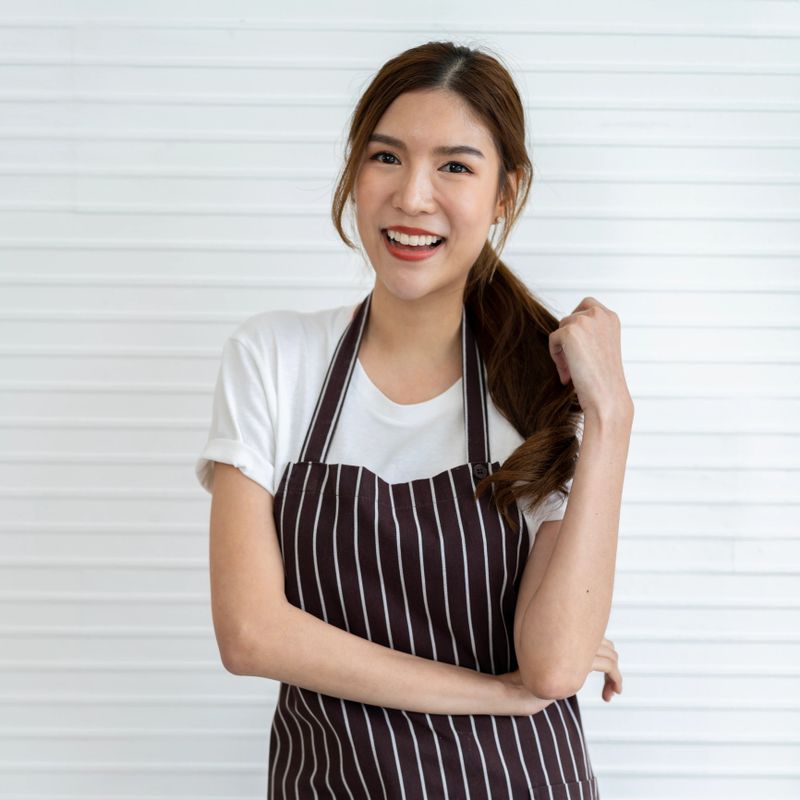A young and beautiful female barista is smiling and posting a stand while wearing a white t-shirt and apron. She is working in a trendy and stylish cafe, serving delicious coffee.