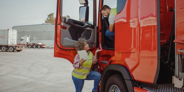 Two workers discussing inside and outside a red truck in a logistics yard.