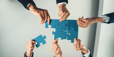 Six hands attempting pull apart large-light blue in color puzzle pieces 