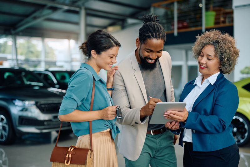 Happy African American man signing a contract on digital tablet while being with his wife on a meeting with car saleswoman in auto salon.