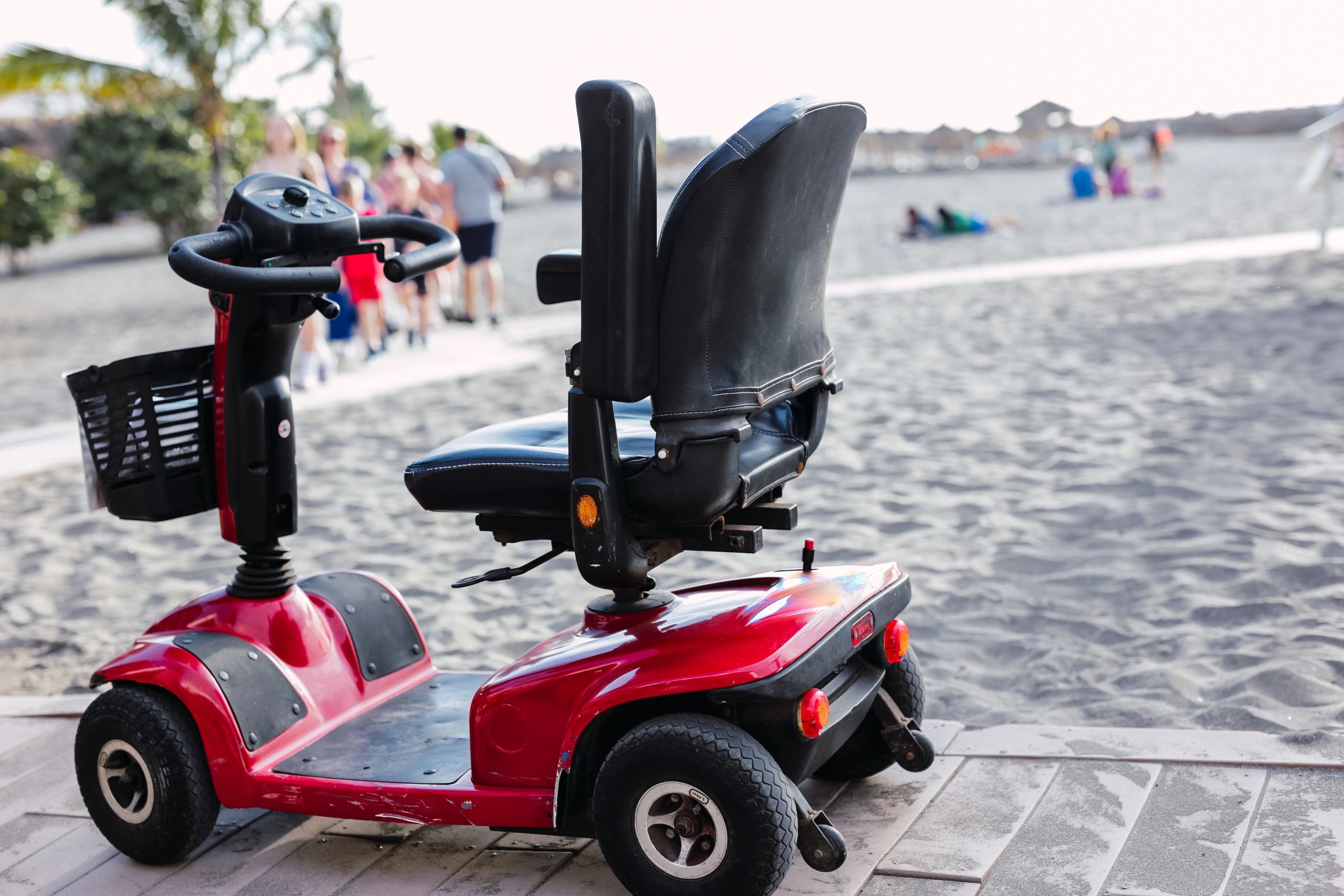 Red mobility scooter parked near a sandy beach with people in the background.