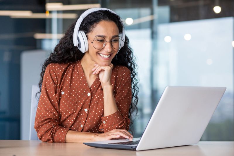 Young beautiful woman in headphones working with laptop inside office at workplace, joyful and satisfied business woman watching online webinar, educational video course, learning remotely.