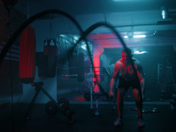 A muscular man exercises with battle ropes in a dimly lit gym.
