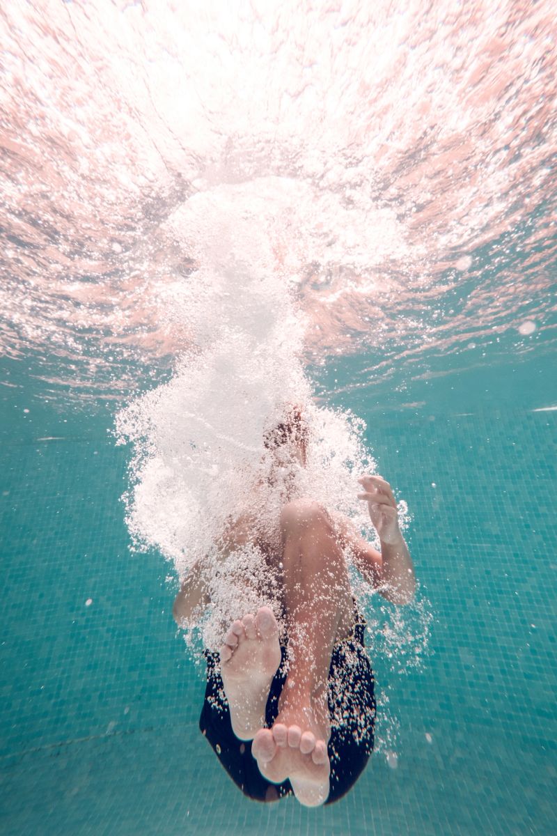 Underwater full body shot of unrecognizable barefoot kid wearing swimming shorts plunging in transparent clean blue water in pool