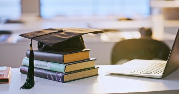 Graduation cap on stack of books beside a laptop on a desk.
