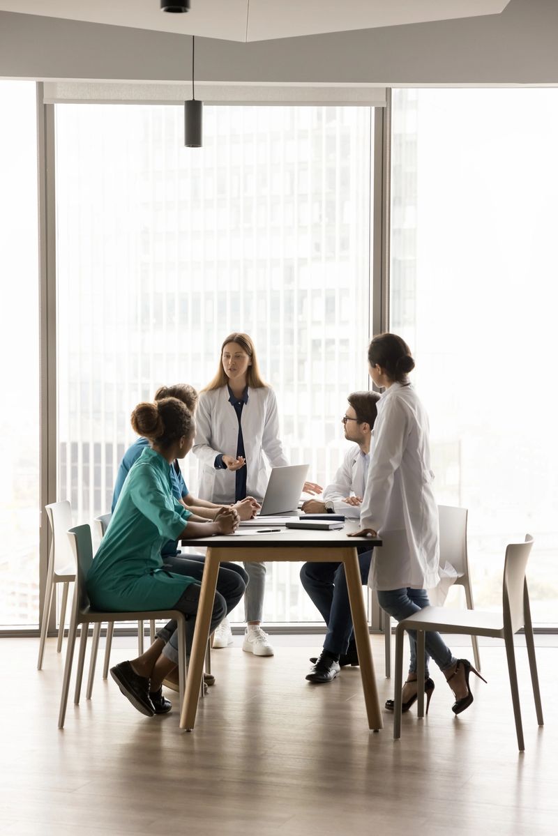 Young female chief doctor holding meeting with diverse medical team in office, standing at large table at boss place, talking to colleagues, instructing staff. Full length wide shot