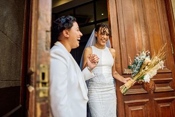 Two brides joyfully holding hands outside a wooden door, one holding a bouquet.