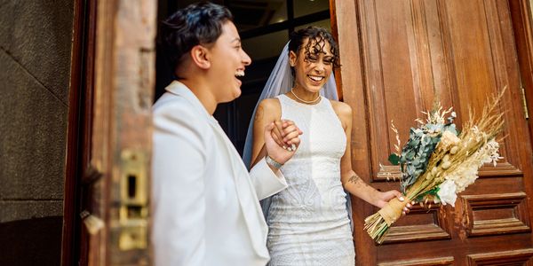 Two brides joyfully holding hands at a wooden door.