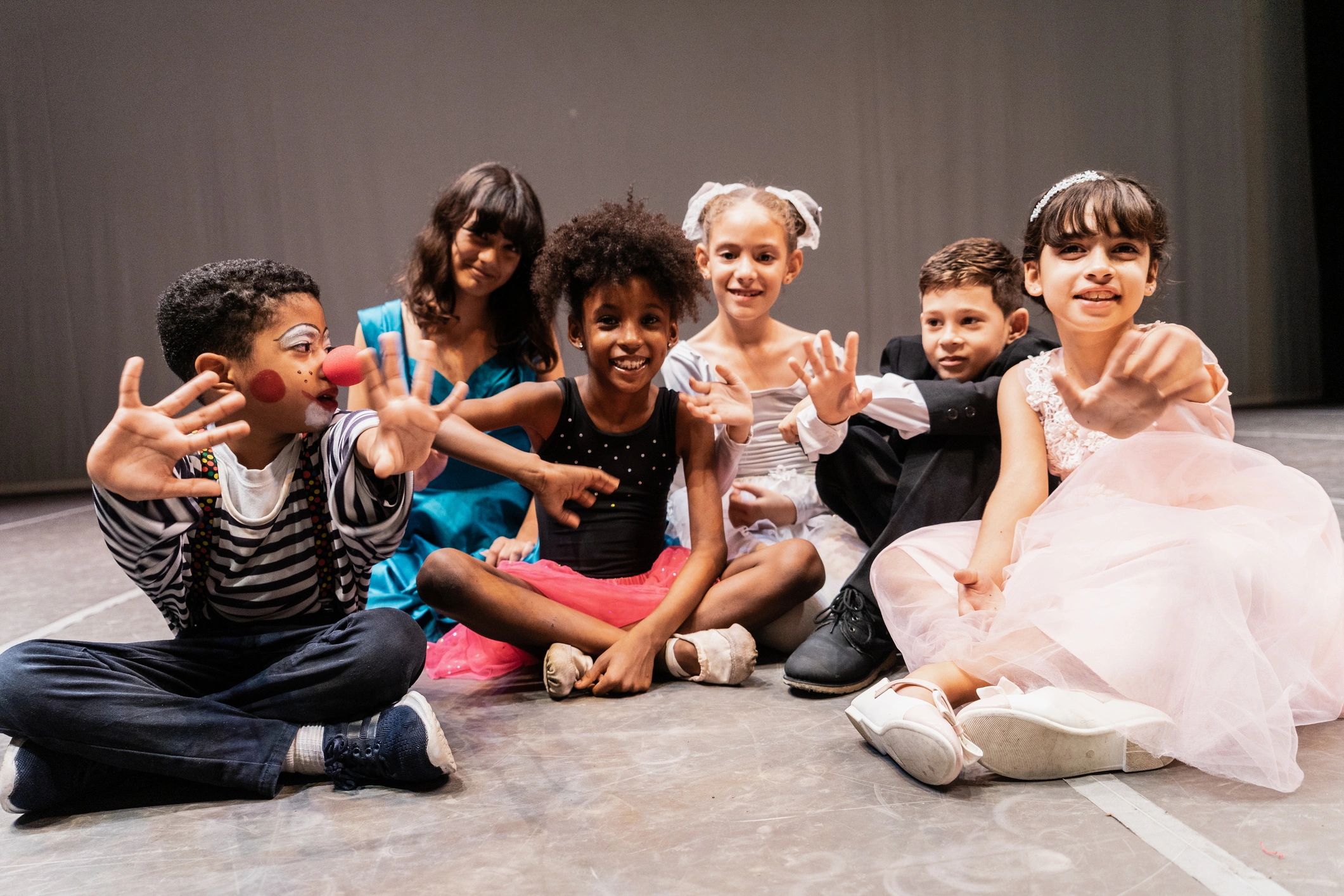 Children in colorful costumes sit together, smiling and waving on a stage.