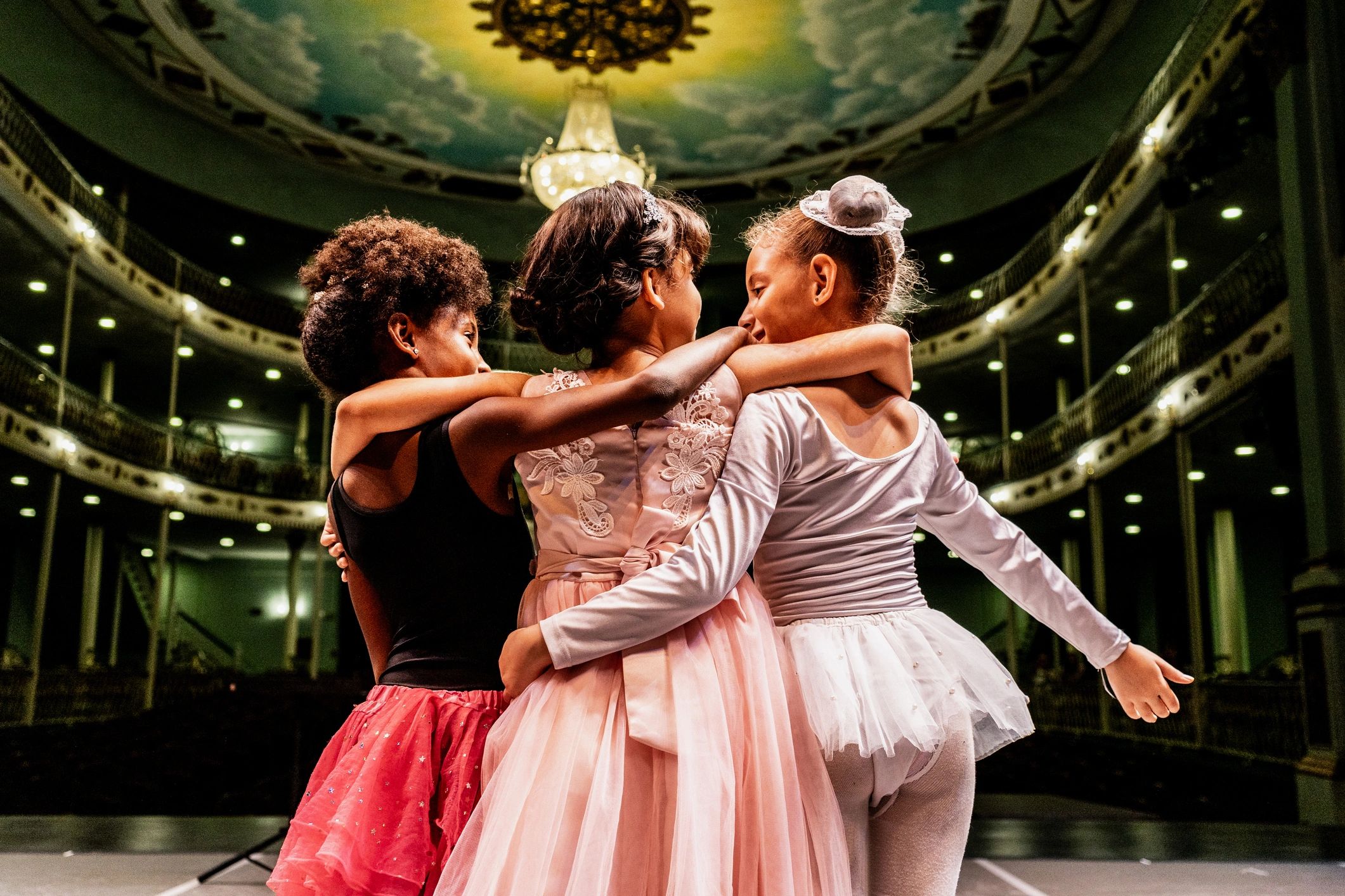 Three young ballerinas embrace on stage in a grand theater.