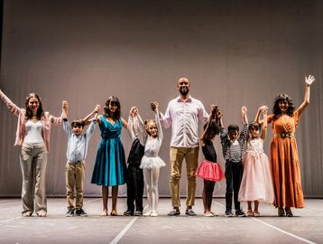A diverse group of performers takes a bow on stage, holding hands and smiling.