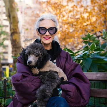 Elderly woman with large sunglasses smiling, holding a fluffy dog in a park.