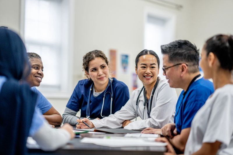 A small medical staff team sit around a boardroom table as they meet to discuss and collaborate on patient cases.  They are each dressed professionally and are focused on the meeting.