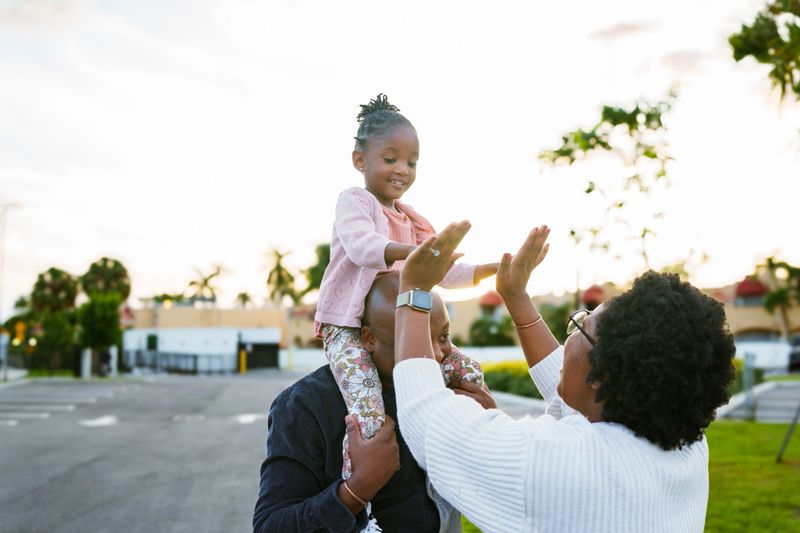 A three year old African American girl has fun riding on her father's shoulders, and gives her mother a high five during a family walk around the neighborhood at sunset.