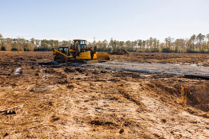 Dirty, puddled area with dry grass. Bulldozer working, making flat plot in the center of the field. Side view of the machinery