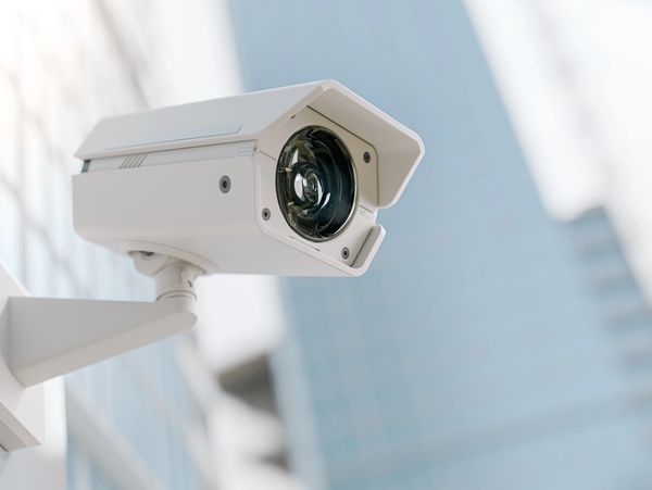 A white surveillance camera mounted on a wall with a blurred cityscape background.