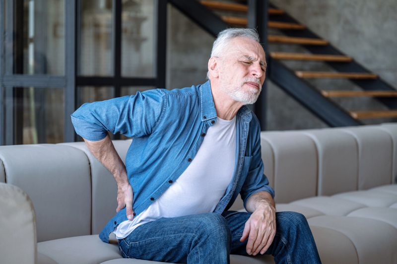 Middle aged man feeling unwell and suffering from chronic backache. Unhappy mature male sitting alone on sofa in living room has discomfort and pain in back. Lumbago symptom
