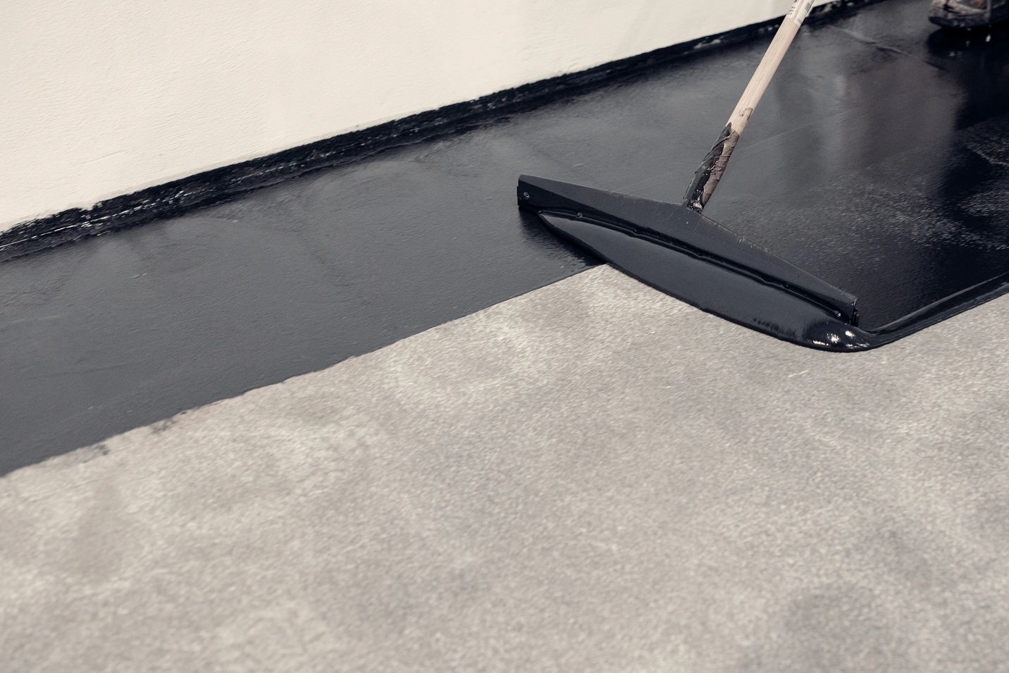 A person applying black waterproof coating on a concrete floor.