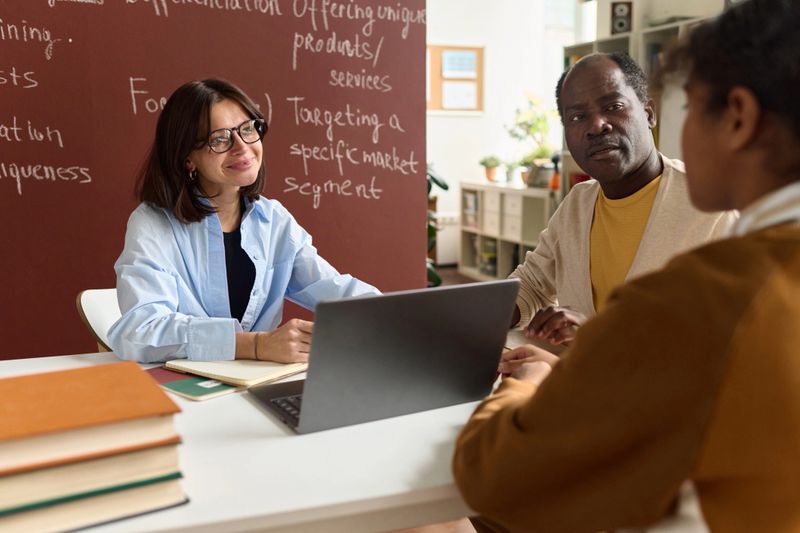 Portrait of smiling female teacher of student advisor talking to girl and parent during meeting in class