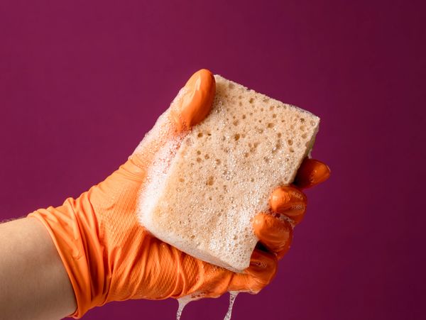 Hand in orange glove squeezing a soapy sponge with foam dripping.