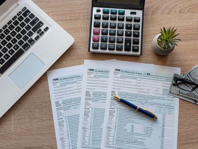 Workspace with tax forms, laptop, calculator, pen, and cash on a wooden desk.